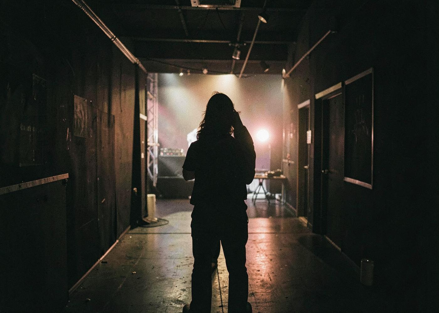Performer silhouetted in a dark backstage corridor with distant stage light